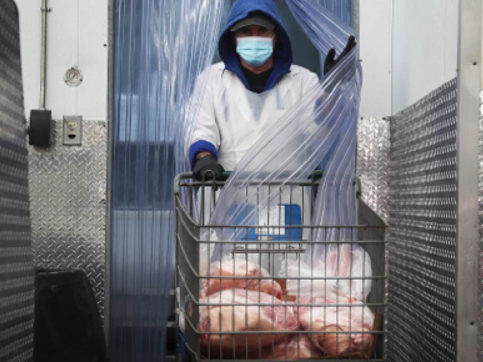 Man wearing a mask and white coveralls pushing a cart with raw meat in a factory setting