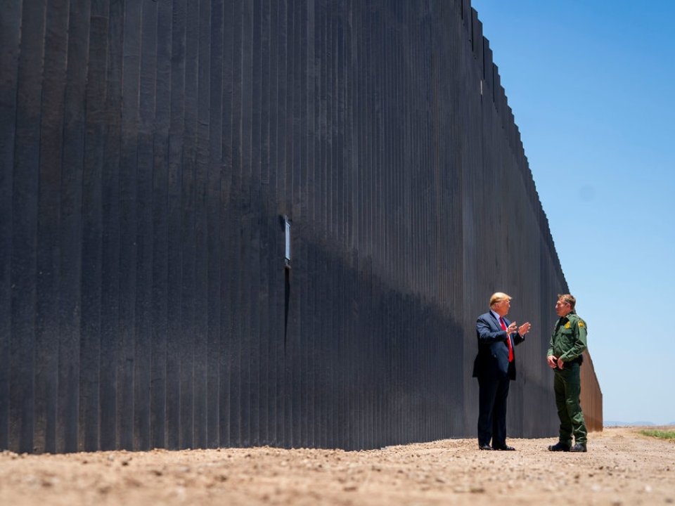 Two men engaged in conversation in front of a wall on a desert-like strip of land