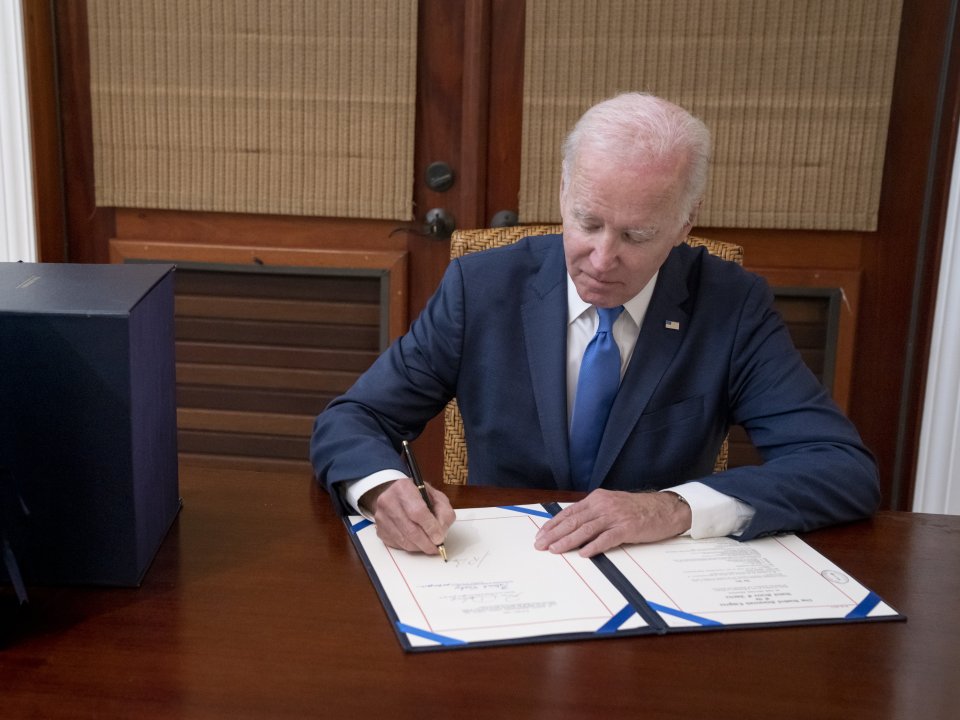 President Biden signs a document while sitting behind a wooden desk.