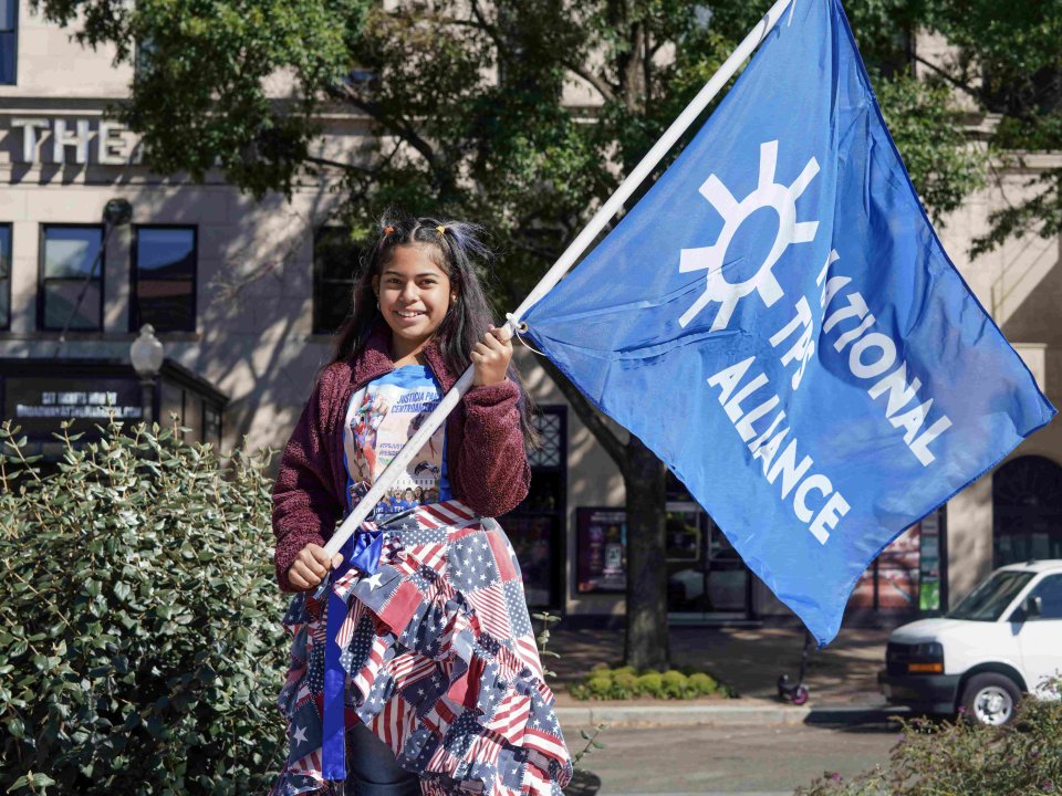 A girl wearing a patterned American flag dress holds a flag that reads "National TPS Alliance."